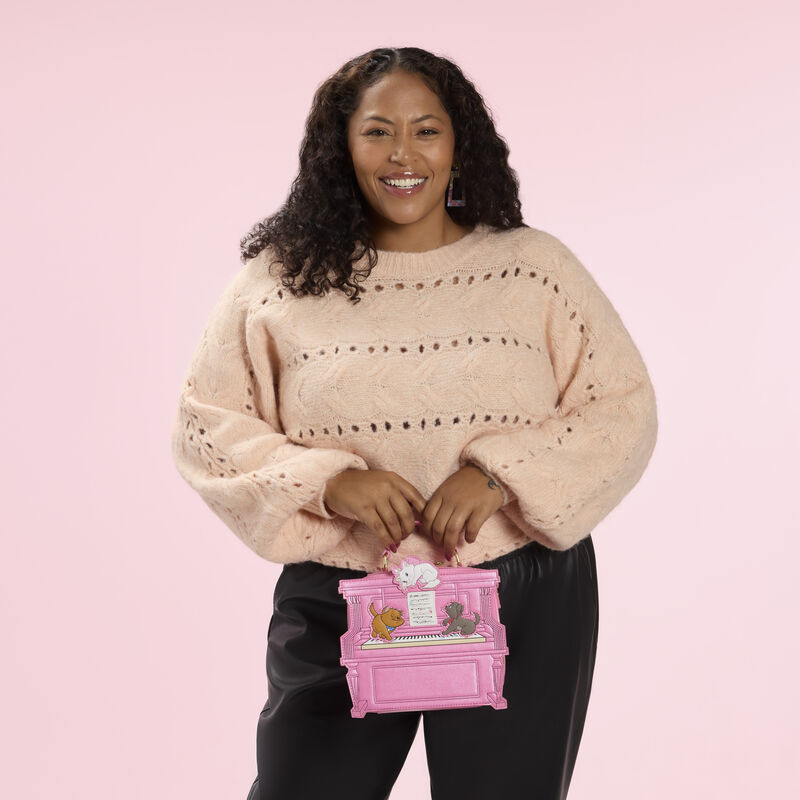 Woman wearing a light pink/beige sweater and black pants. She holds the Loungefly Aristocats Figural Piano Crossbody Bag by the top handle. She stands against a pink background. 