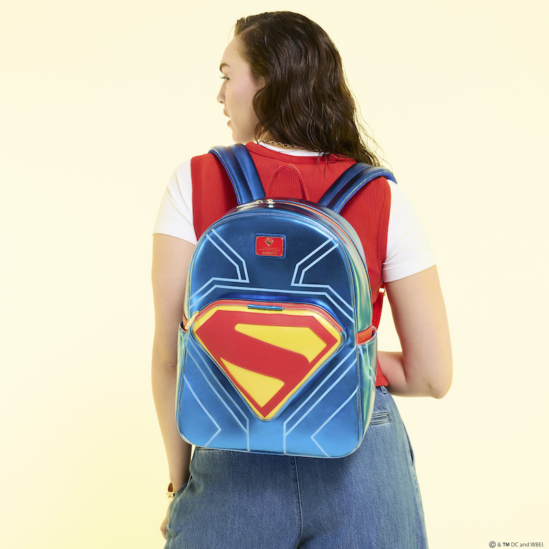 Woman facing away from camera wearing a white-and-red top with blue jeans. She wears the Loungefly Superman Full-Size Light Up Glow Backpack on her back. She stands against a light yellow background
