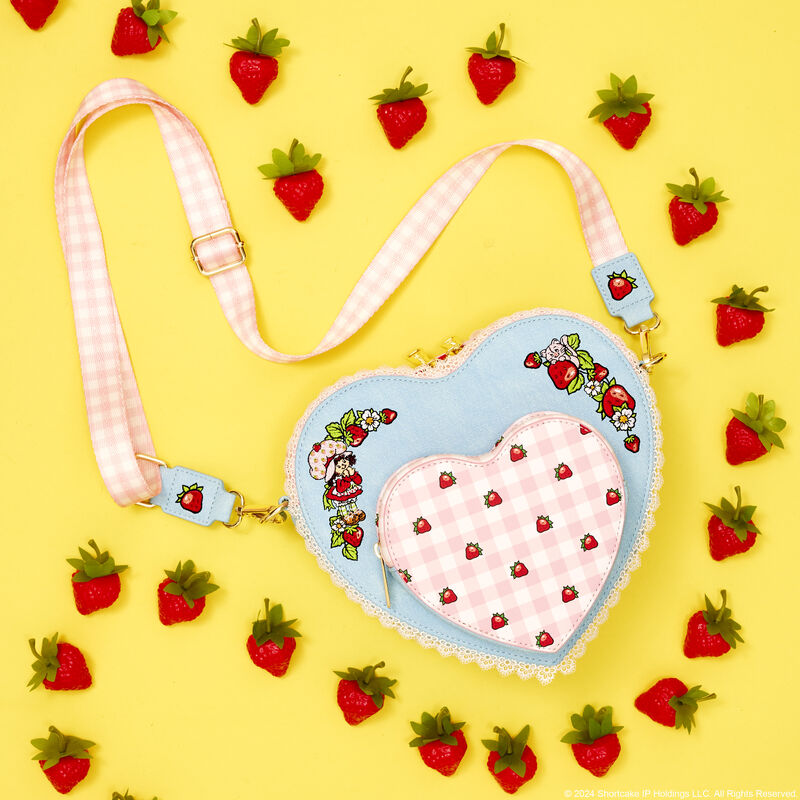 Heart-shaped Loungefly Strawberry Shortcake crossbody bag laying against a yellow background surrounded by strawberries