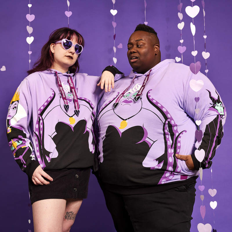 Image of a woman and man standing together and wearing the Loungefly Disney Villains Curse Your Hearts Unisex Hoodie, standing against a purple background with purple hearts raining down.