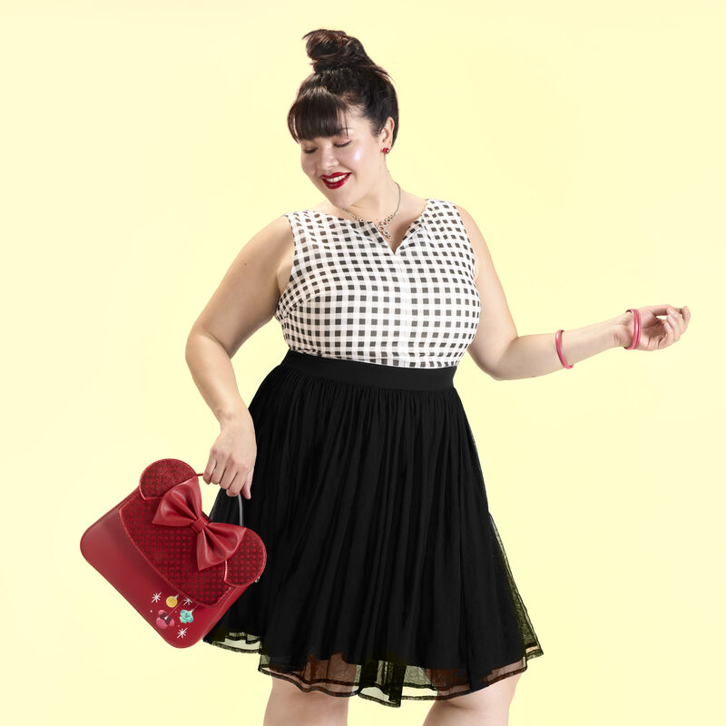 Woman wearing a white checkered blouse and black skirt, holding the Loungefly Minnie Mouse holiday crossbody bag. She stands against a yellow background.