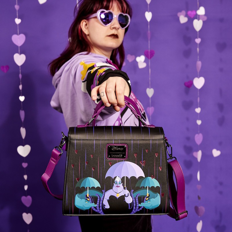 Image of woman holding the Disney Loungefly Disney Villains Curse Your Hearts Crossbody Bag, showing off the back toward camera to show off the artwork of Ursula with Flotsam and Jetsam holding umbrellas in the rain 