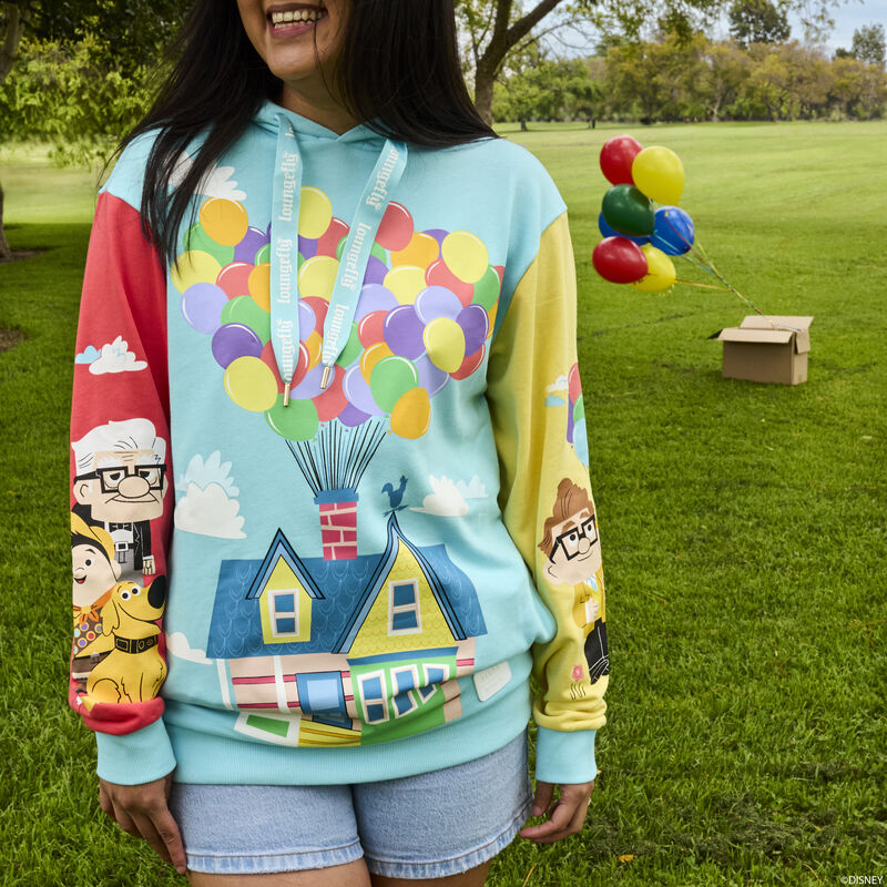 Woman with long dark hair standing outside in a park wearing the Loungefly Pixar Up 15th Anniversary Color Block Hoodie, featuring Carl and Ellie's house on the torso against a blue background; Carl, Russel, and Dug on the red sleeve; and a young Carl and Ellie on the yellow sleeve.