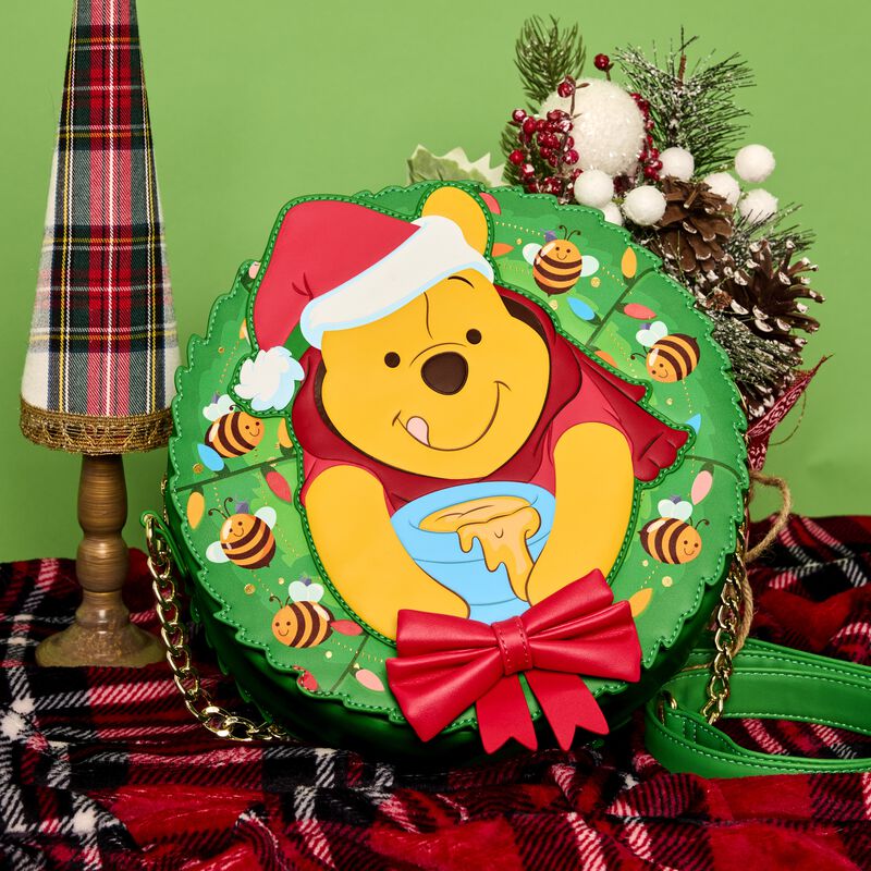 Loungefly crossbody bag featuring Winnie the Pooh sticking out from the middle of a holiday wreath. The bag sits on a table covered in a plaid table cloth and is surrounded by holiday decor, like pine cones. 