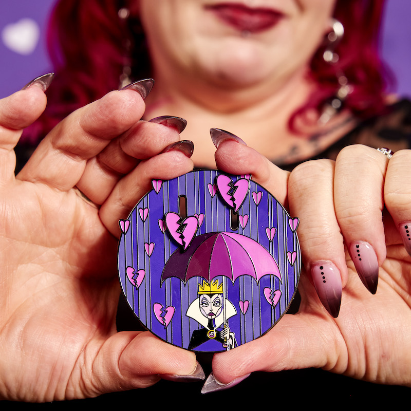 Image of woman holding the Loungefly Disney Villains Curse Your Hearts 3" Collector Box Sliding Pin out to the camera, showing the Evil Queen holding an umbrella while purple hearts rain down on her