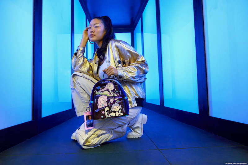 Woman wearing white cargo pants, white boots, a white crop top, and a metallic gold jacket. She knees down and holds the Loungefly KPop Demon Hunters HUNTR/X mini backpack. Behind her is a blue hallway featuring light blue lights. 