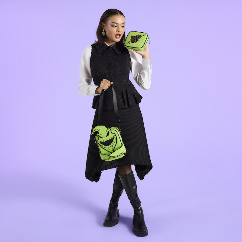 Woman wearing a black vest over a white long-sleeved top with a black skirt and knee-high boots. She holds the Oogie Boogie Crossbuddies bag by the strap in one hand and holds up the coin pouch in the other. She stands against a purple background. 
