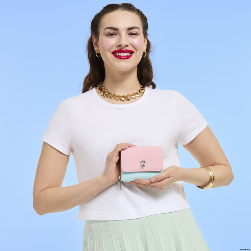 Woman smiling at camera wearing a white top and a green skirt. She holds the Loungefly Classic Barbie wallet in her hands. She stands against a blue background. 
