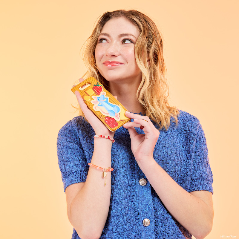 Woman looking up to the left and holding the Loungefly Ratatouille Charcuterie Card Holder. She smiles and wears a blue button-down sweater. She stands against a peachy background. 