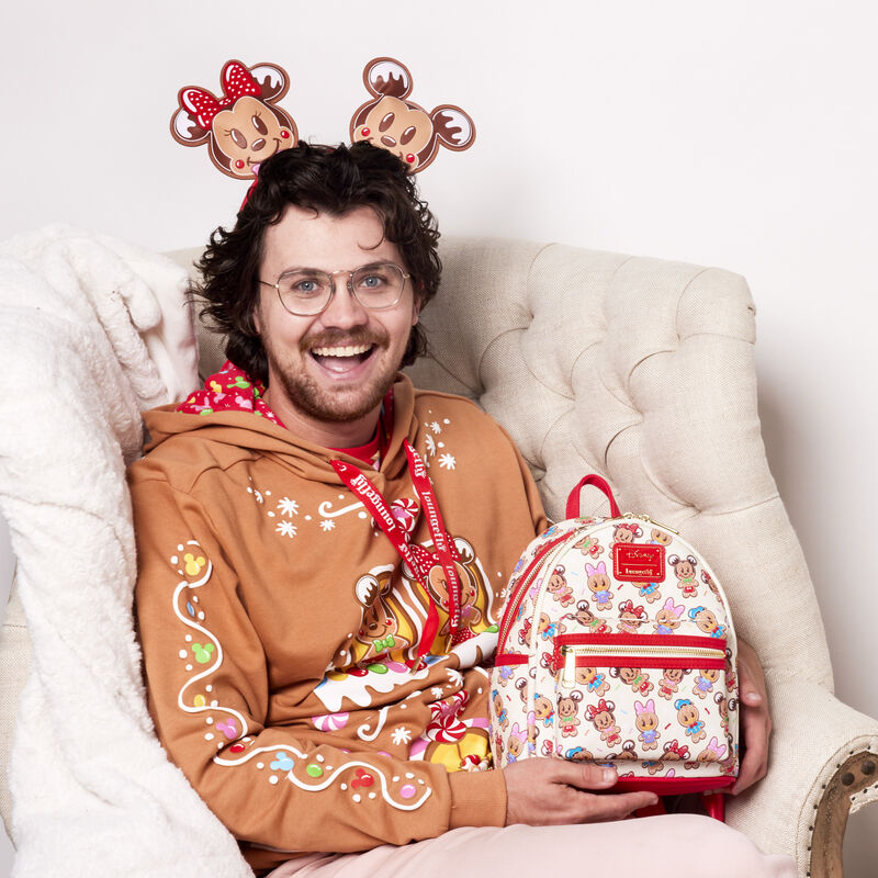 Man sitting in a white chair wearing the Mickey and Friends Gingerbread House Unisex Hoodie and holding the Mickey and Friends Gingerbread Cookie All-Over Print Mini Backpack and wearing the matching ear headband.