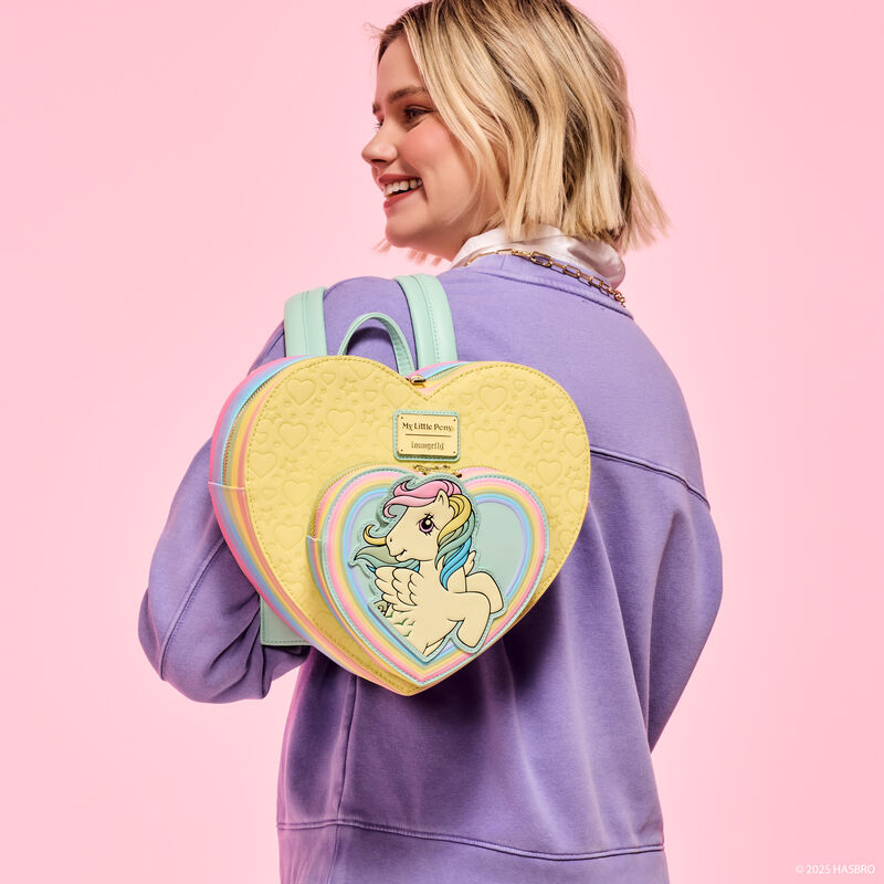 Woman wearing a purple sweatshirt facing away from camera and smiling over a shoulder. She wears the heart-shaped Loungefly My Little Pony Skydancer mini backpack. She stands against a pink background. 