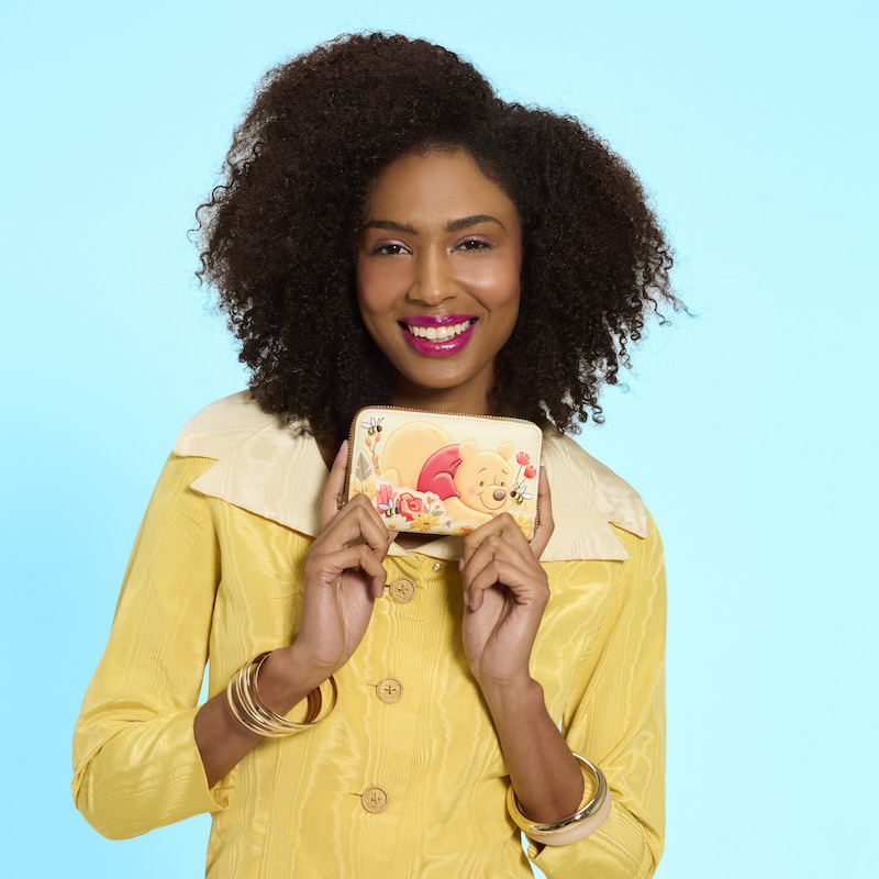 Woman wearing a yellow dress, holding up the Loungefly Winnie the Pooh wallet up and smiling at camera. She stands against a blue background. 