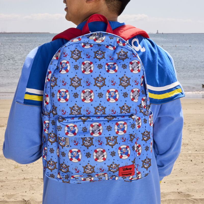 Man at the beach wearing the Loungefly Donald Duck 90th Anniversary Hoodie and Full-Size nylon backpack, featuring an all-over print of Donald Duck and other sailing motifs