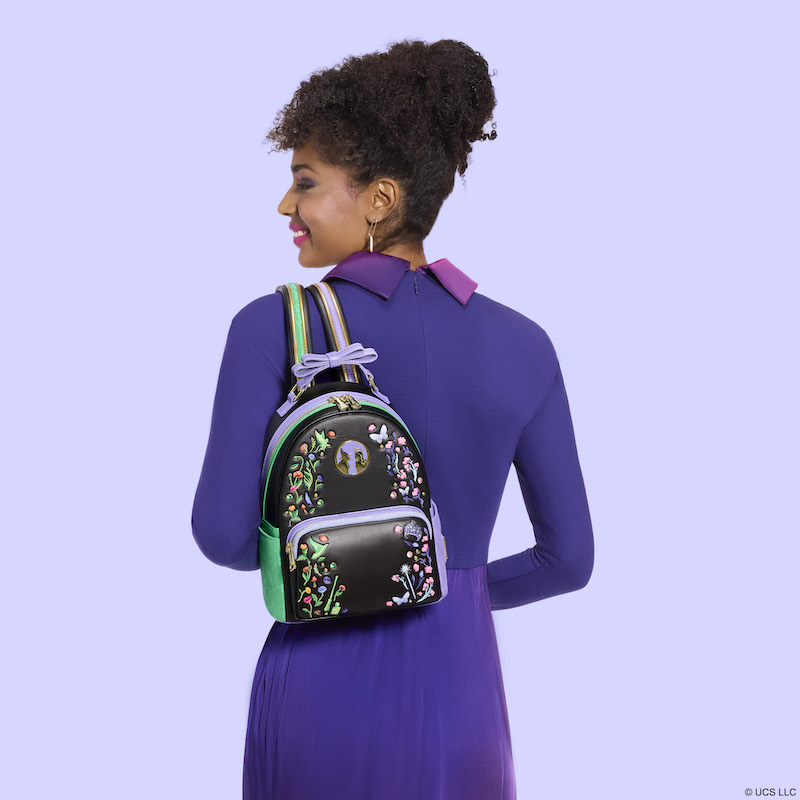 Woman wearing a purple dress, facing away from camera. She smiles over her shoulder. She wears the Loungefly Wicked Elphaba & Glinda Floral and Lace Mini Backpack over a shoulder. She stands against a purple background. 