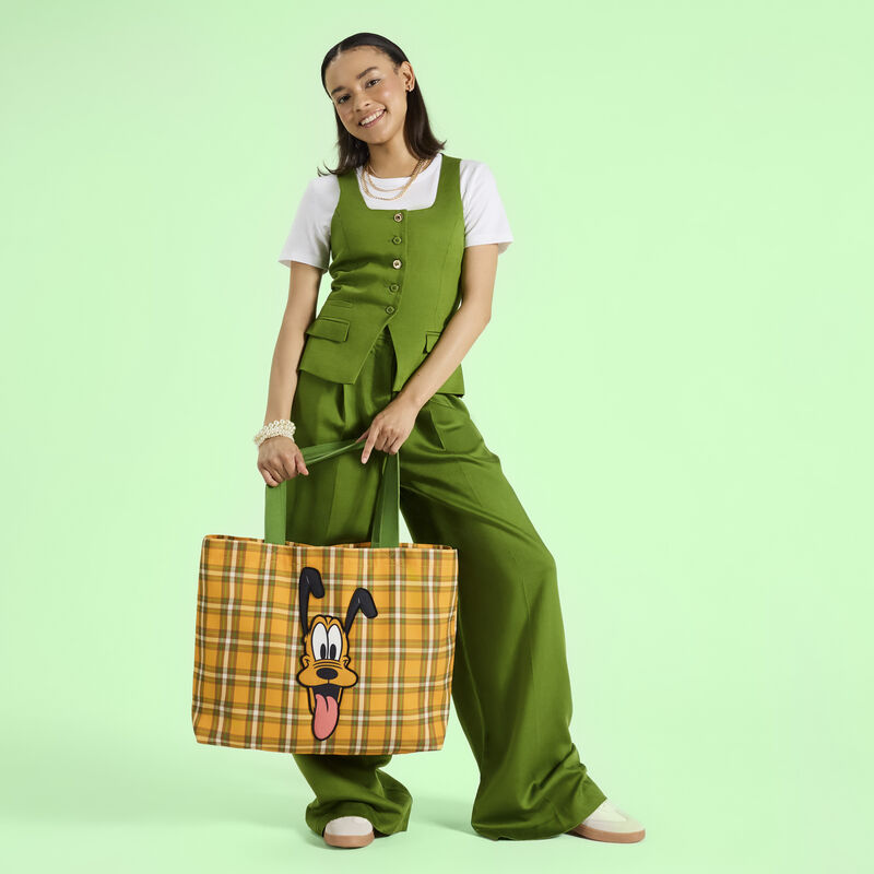 Woman wearing a green vest over a white top and green trousers, posing with the Loungefly Pluto 95th anniversary canvas tote. She stands in front of a green background. 