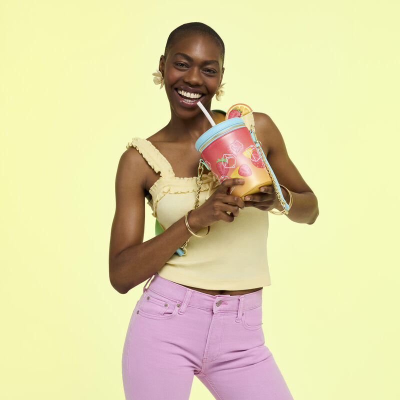Woman smiling at camera holding up the Loungefly Tropical Drink Crossbody. She wears a yellow tank top and pink jeans and stands in front of a yellow background. 
