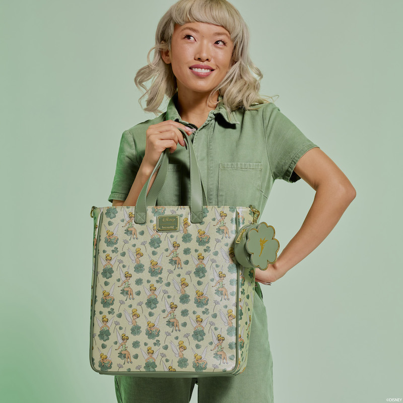 Woman wearing a green jumpsuit smiling and look up, holding the Loungefly Peter Pan Tinker Bell Clover Tote Bag. She stands against a light green background. 