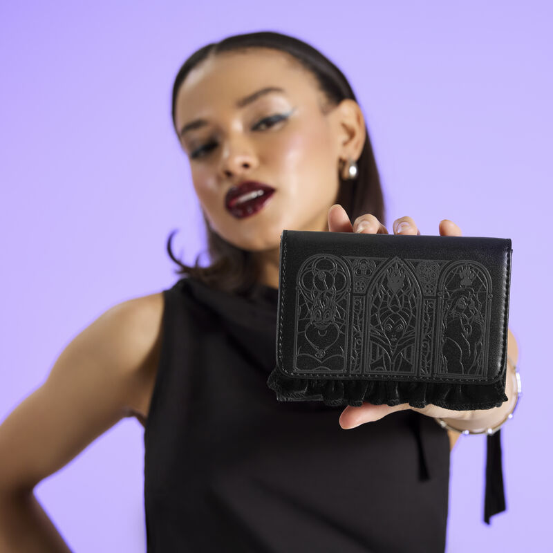 Woman wearing a black top, holding the Loungefly Disney Villains Stained Glass Trifold Wallet out to camera. She stands in front of a purple background. 
