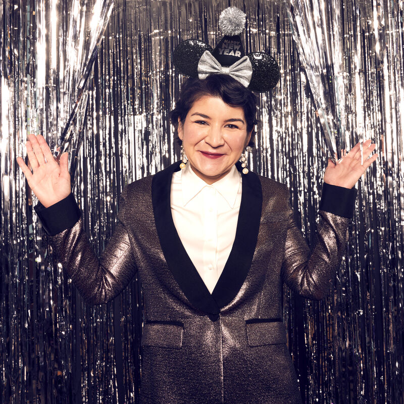 Image of person standing in front of silver streamers in silver blazer wearing the Minnie Mouse Happy New Year Black Glitter Ear Headband 