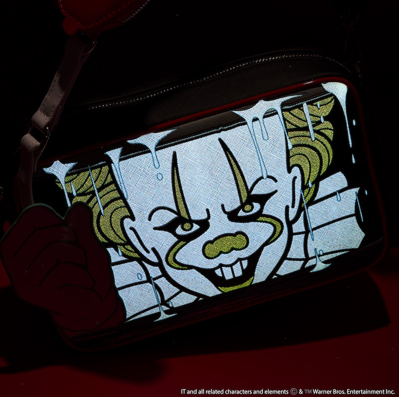 Loungefly Pennywise It Glow Crossbody bag against a dark background showing off the glow in the dark features of Pennywise and the rain.