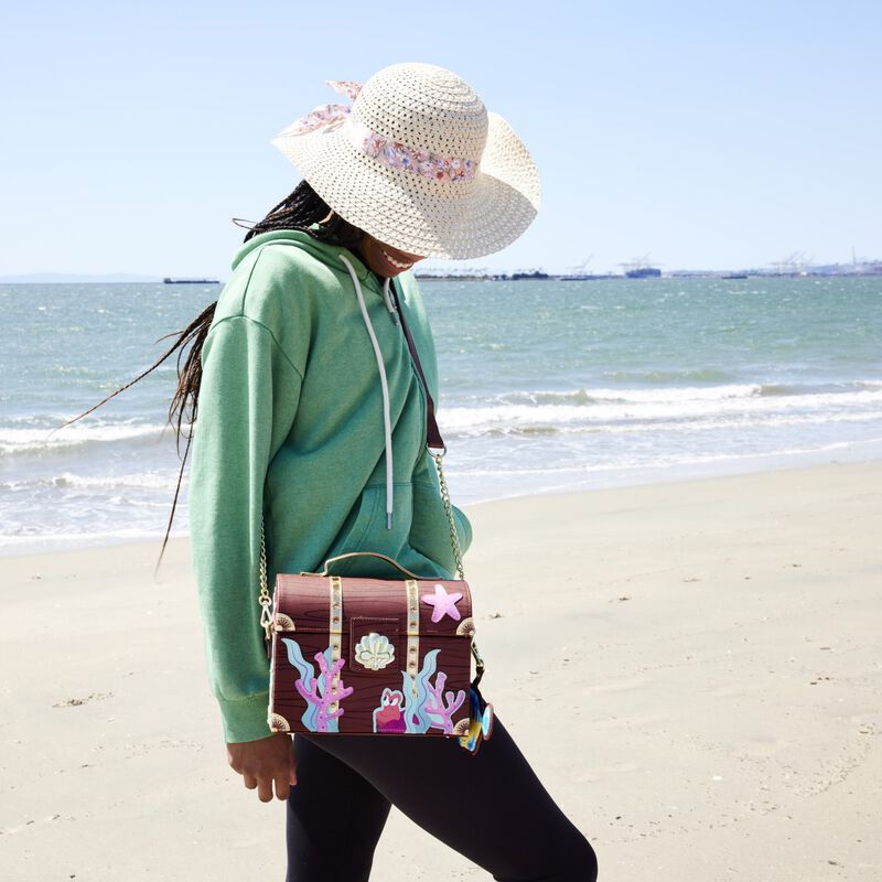 Woman at the beach wearing a green hoodie and white hat wearing the Little Mermaid Treasure Chest Crossbody