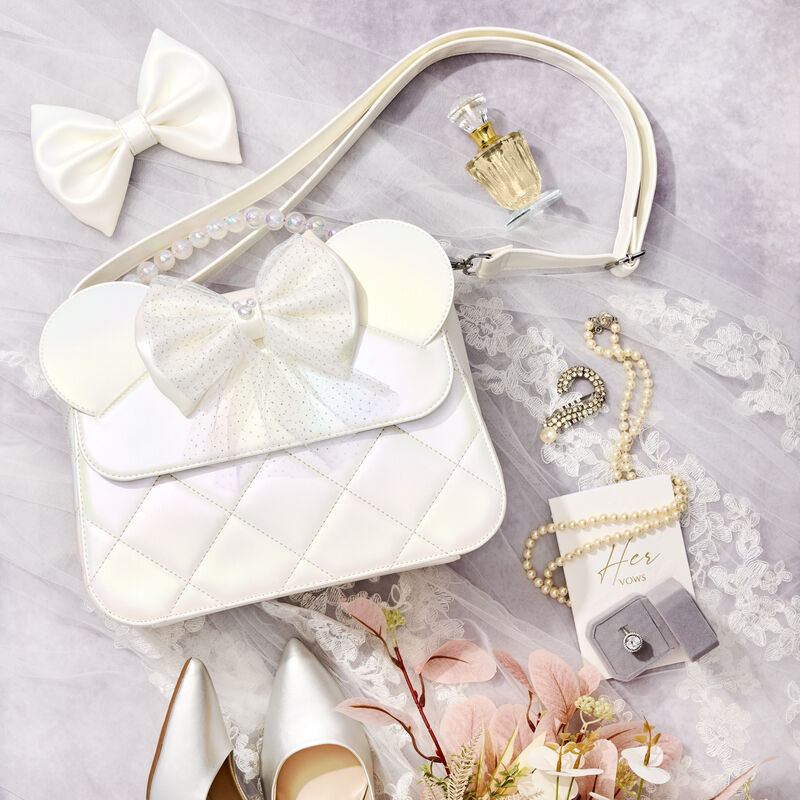 Flat lay of the Loungefly Disney Minnie Mouse Iridescent Wedding Crossbody Bag laying against a veil and surrounded by other wedding accessories including a perfume bottle, a pearl necklace, a bow, and silver heels.