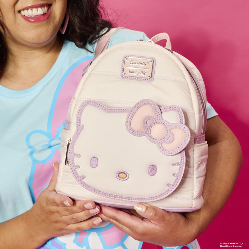 Close-up shot of woman smiling at camera and holding the Loungefly Sanrio Hello Kitty Quilted Texture Mini Backpack, featuring Hello Kitty on the front pocket outlined in purple with white quilted material on the bag.