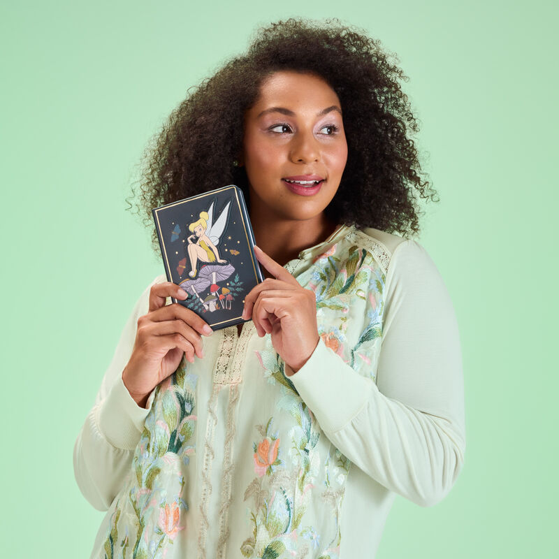 Woman wearing a green floral blouse, holding the Tinker Bell journal and standing against a green background. 