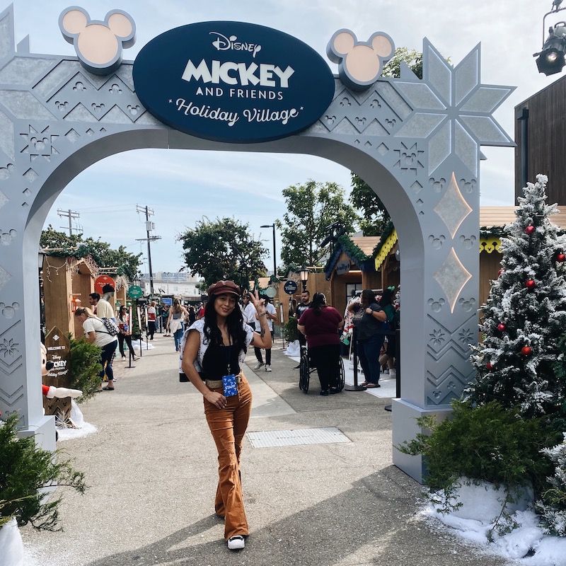 Woman standing under an archway beneath a sign that says "Disney Mickey and Friends Holiday Village" smiling and throwing up a peace sign with her hand