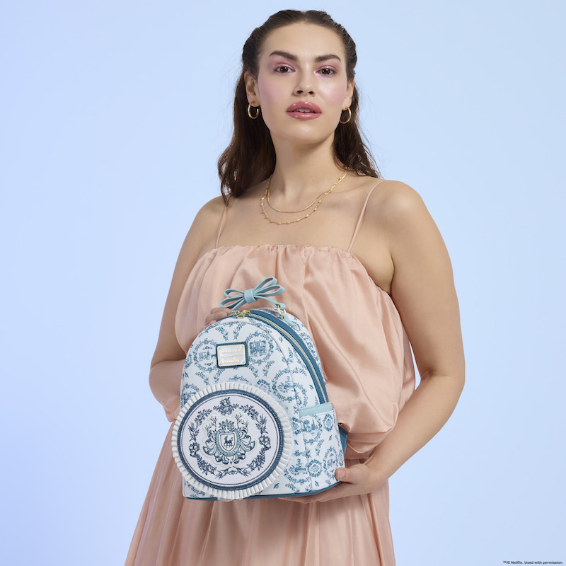 Woman standing in front of a light blue background wearing a pink silk dress, holding the exclusive Loungefly Bridgerton mini backpack.  