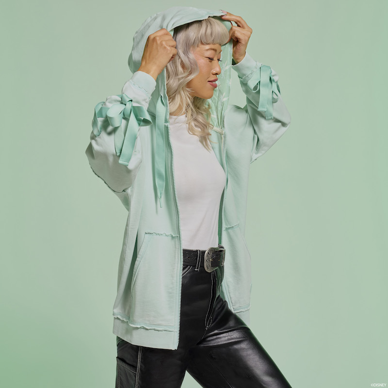 Woman faces to the right and wears the light green Loungefly Peter Pan Tinker Bell Ribbon Zip Up Hoodie. The hoodie is unzipped over a white shirt and black jeans. The woman pulls the hood over her head. She stands against a light green background. 