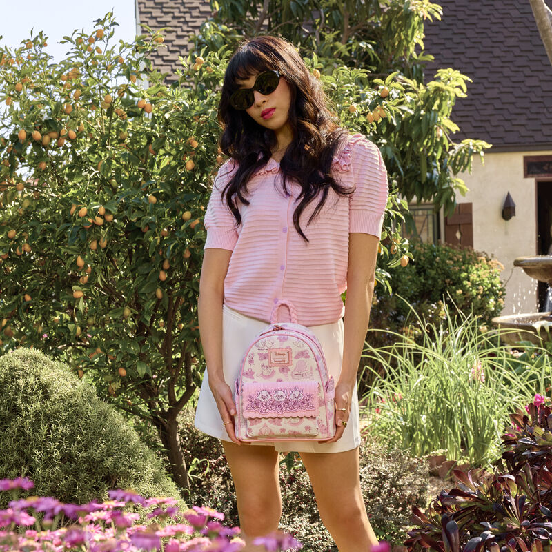 Woman standing outside wearing a pink top and white skirt. She holds the Loungefly The Aristocats all-over print mini backpack. 