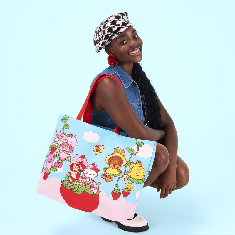 Woman squatting and posing against a light blue background wearing a denim vest and black-and-white houndstooth hat. She wears the Loungefly Sanrio Hello Kitty & Friends x Strawberry Shortcake tote bag. 