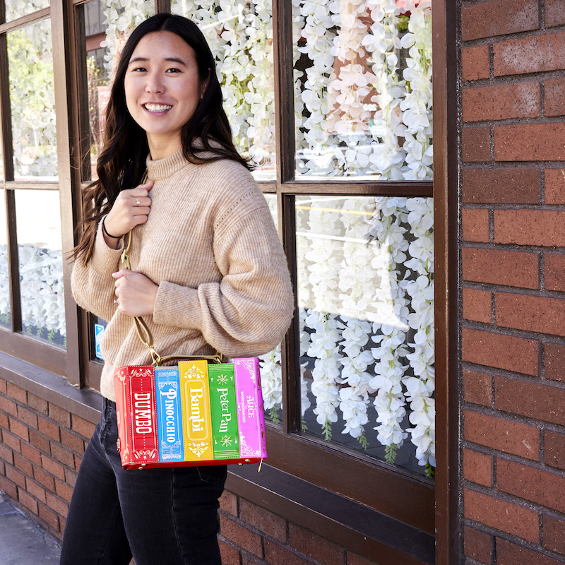 Woman outside against a brick wall and a window with flowers in it, wearing the Stitch Shoppe Classic Disney Books Crossbody Bag