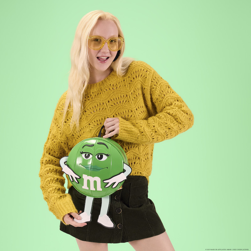 Woman wearing a yellow sweater and black skirt holding the Loungefly x M&M’S Green Character mini backpack.