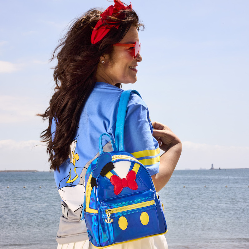 Woman standing at the beach with a red bow in her hair, facing away from camera and wearing the metallic Donald Duck 90th Exclusive Mini Backpack 