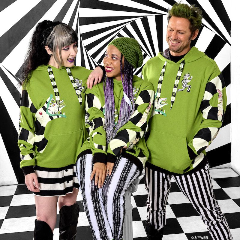 A man and a woman standing beside a woman who's sitting, all three of them wear the black, white, and green Loungefly Beetlejuice Unisex Hoodie. They're smiling at each other and standing against a black and white background.