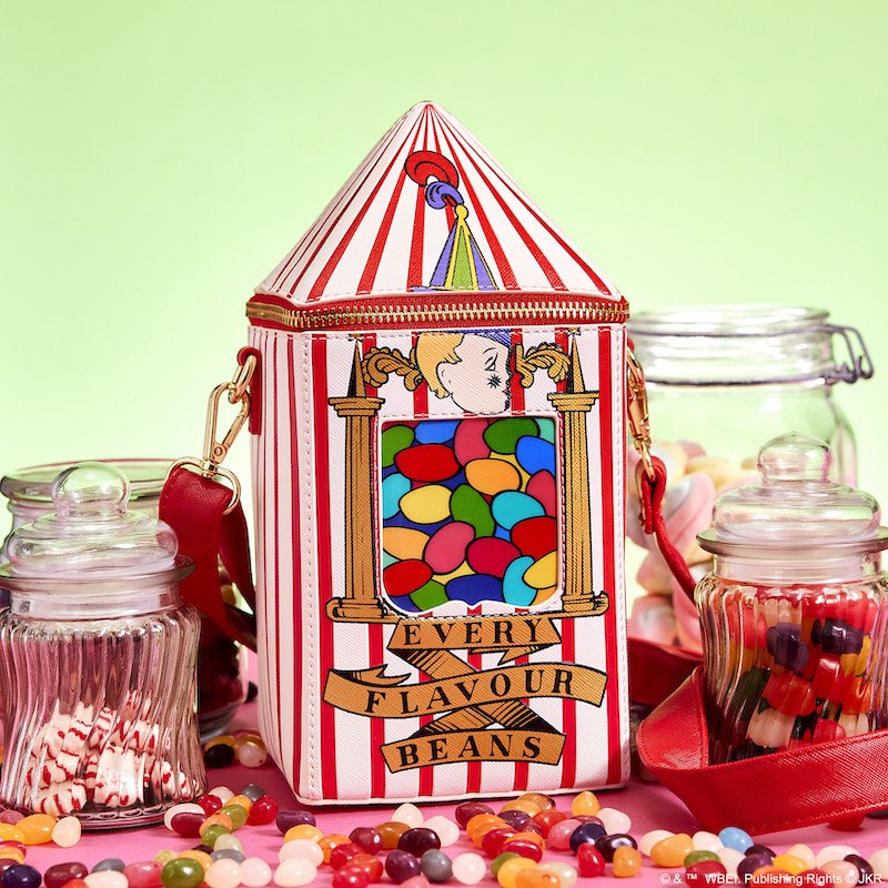 Red and white figural crossbody bag in the shape of a container of Bertie Bott's Every Flavour Beans on a table surrounded by candy