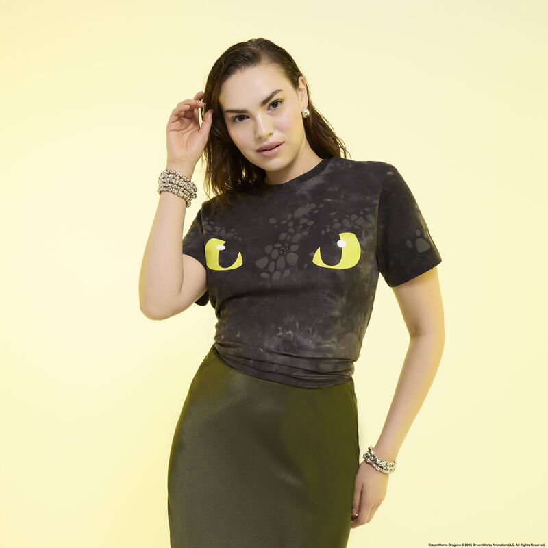 Woman posing in front of camera wearing the black Loungefly Night Fury Toothless Unisex Tee and a green skirt. She stands against a light yellow background. 