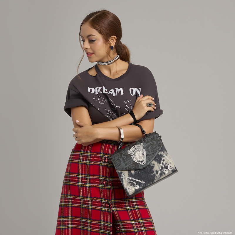 Woman wearing red plait skirt and black t-shirt that says “Dream On”, holding the Loungefly Stranger Things Hellfire Club Crossbody Bag over an arm. She stands against a gray background. 