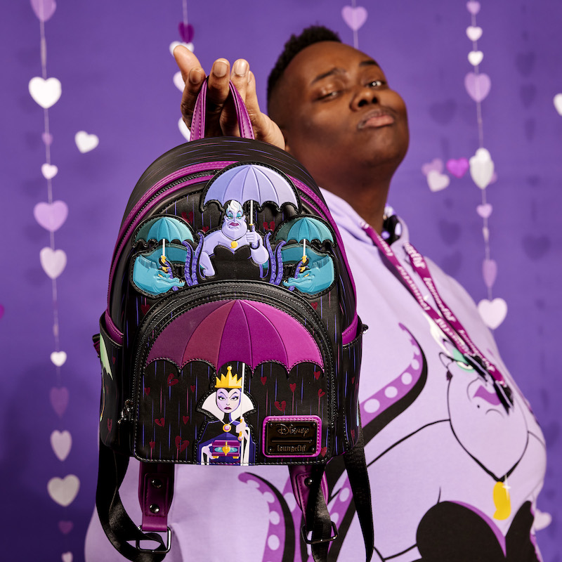 Image of man holding the Loungefly Disney Curse Your Hearts Mini Backpack toward the camera while wearing the Curse Your Hearts Hoodie and standing against a purple background with Purple Hearts raining down 