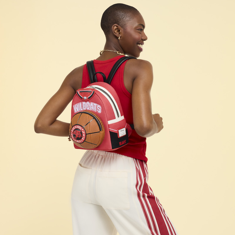 Woman facing away from camera wearing a red top and white pants with red stripes. She wears the Loungefly High School Musical Troy Bolton Wildcats Jersey mini backpack on her back and stands in front of a yellow background. 