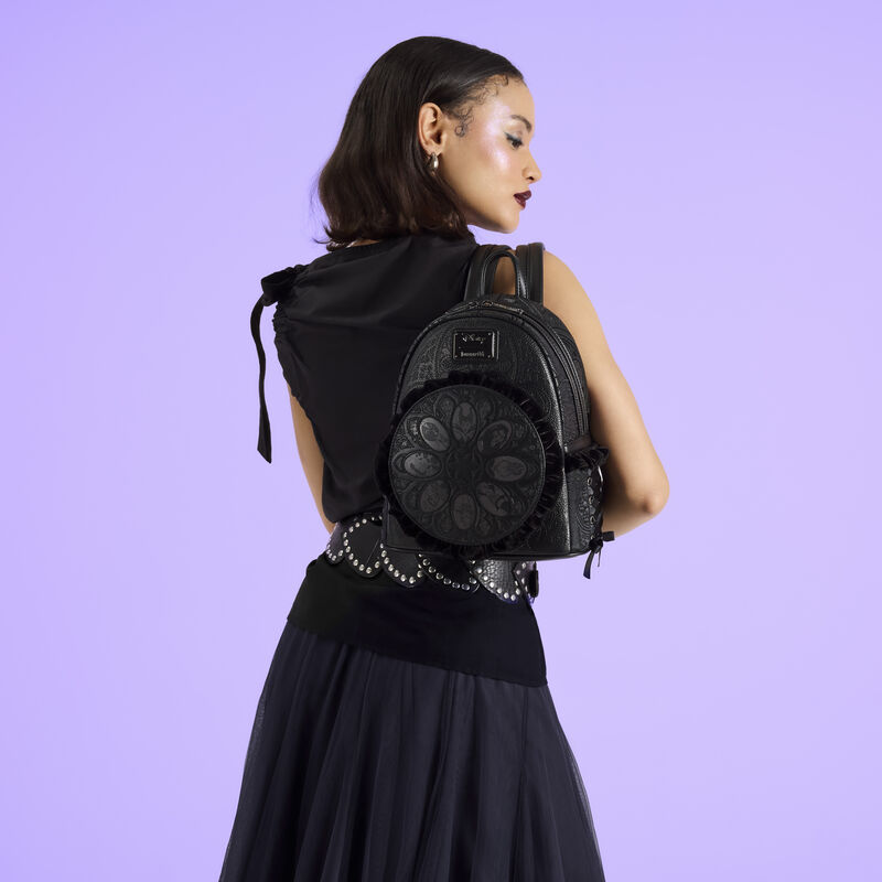 Woman facing away from camera wearing a black top, belt, and skirt. She wears the Loungefly Disney Villains Stained Glass mini backpack over a shoulder. She stands in front of a purple background. 
