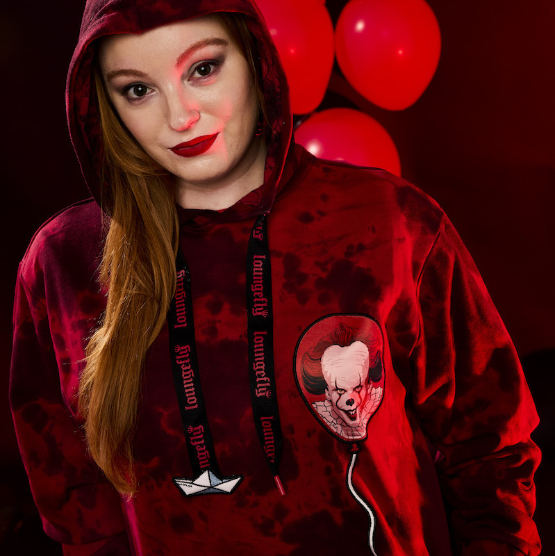 Woman standing against a dark background with red balloons, wearing the It hoodie that features black-and-red tie dye and Pennywise in a balloon on the torso. 