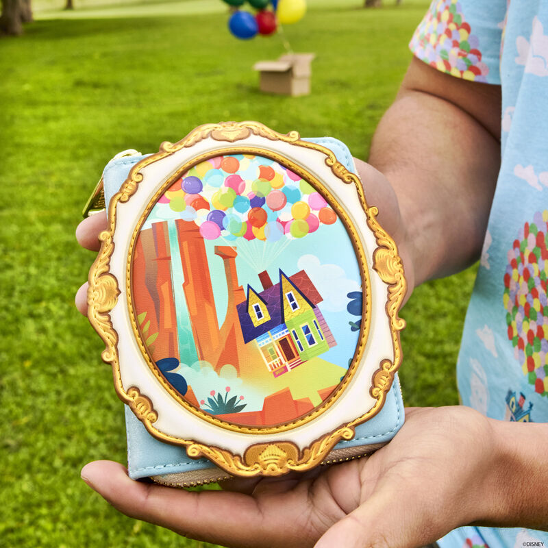 Close up image of a person standing outside in a park holding out the Loungefly Pixar Up Exclusive 15th Anniversary Carl and Ellie Cameo Zip Around Wallet to the camera, showing an image of their house floating at Paradise Falls within a gilded frame.