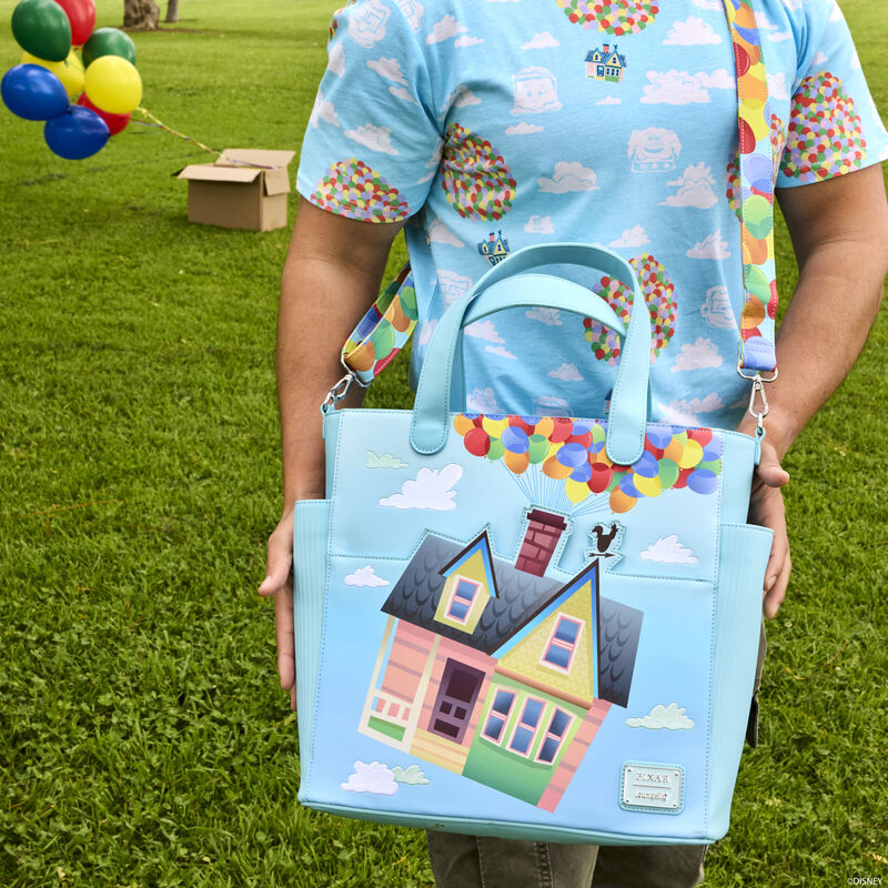 Person standing outside in a park with green grass wearing the Loungefly Pixar Up 15th Anniversary Balloon House Convertible Backpack & Tote Bag, which is light blue and features Carl and Ellie's house suspended by balloons in the sky. 