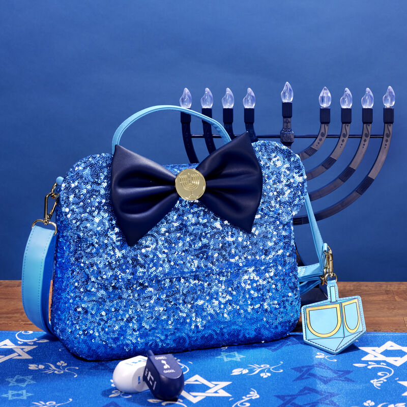 Image of our blue sequin Hanukkah crossbody bag sitting on a table on top of a Hanukkah-themed table runner with a menorah and two dreidels