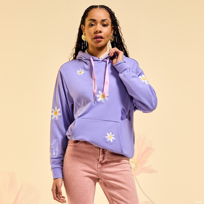 Woman posing in front of camera, wearing the purple Loungefly Daisy Duck 85th Anniversary Hoodie with a pair of light brick-colored jeans. She stands against a beige background. 