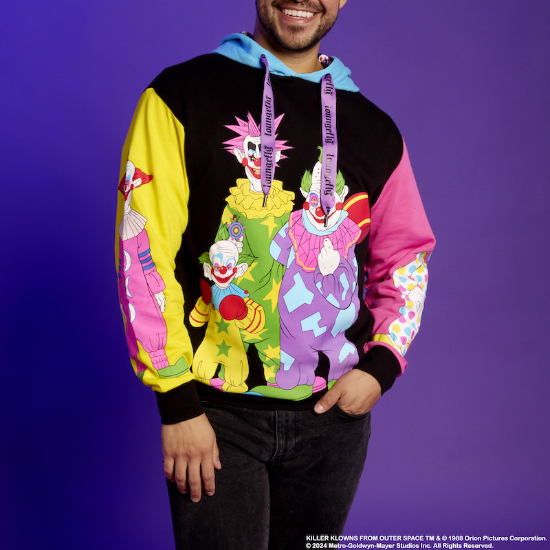 Man standing in front of a purple background wearing the black, pink, yellow, and blue color-blocked Killer Klowns hoodie, featuring the Killer Klowns from Outer Space on the front and sleeves. 