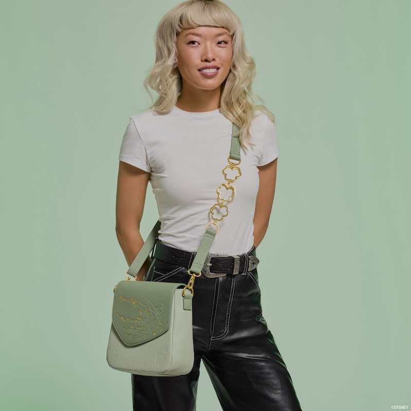 Woman smiling at camera wearing a white shirt and black jeans and wearing the Loungefly Peter Pan Tinker Bell Clover Crossbody bag. She stands against a light green background. 